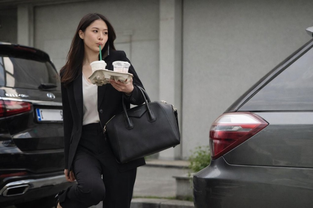 都會女性手持飲品與提袋，穿梭於停車場與車輛之間，象徵通勤接送與多工節奏下的忙碌日常